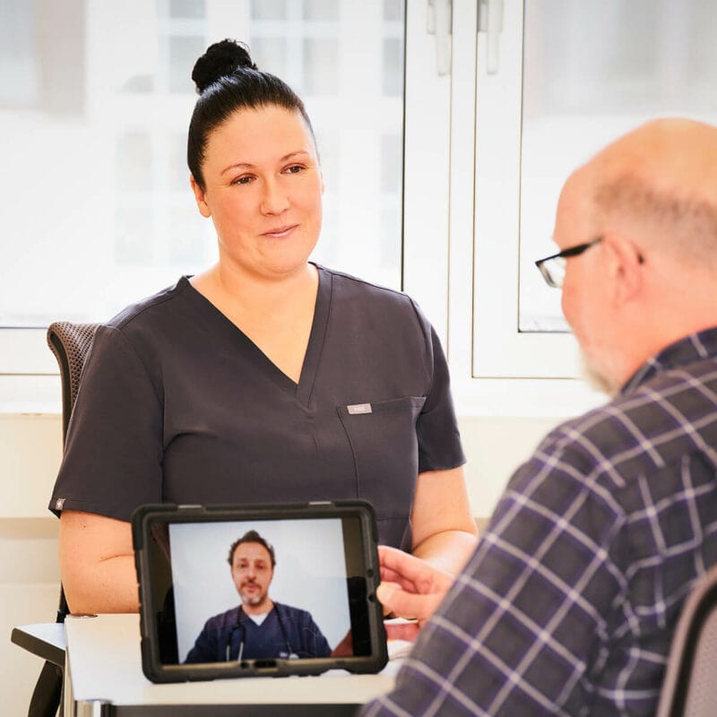 Physician Assistant in einer Lillian-Care-Praxis im Patientengespräch, die angeschlossene Ärztin ist per Tablet zugeschaltet